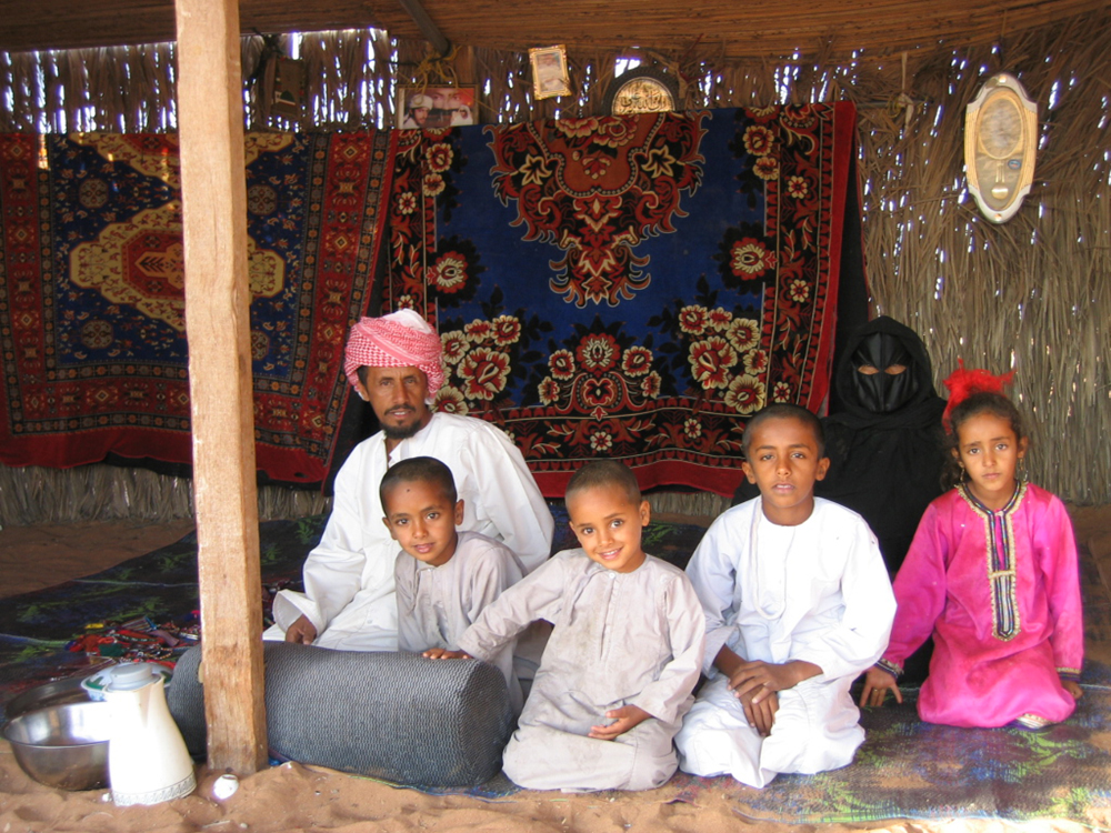 A man in a turban, a woman in a veil covering her face, and four children are shown sitting in a thatched hut. Carpets can be seen on the sandy ground and on the walls of the hut.
