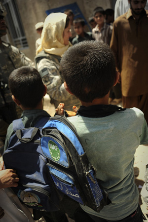 Two Afghan boys with backpacks look toward a smiling U.S. woman in camouflage uniform and yellow headscarf. 