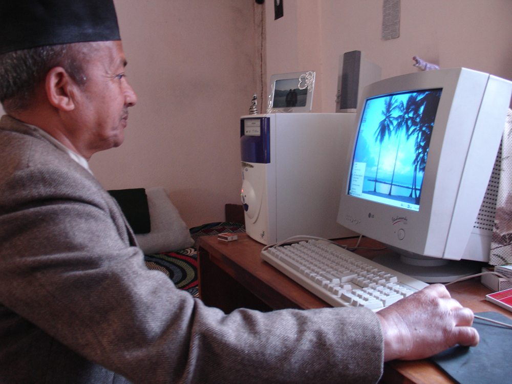 In figure (b), a man wearing a sports jacket and a hat is shown working on a computer.