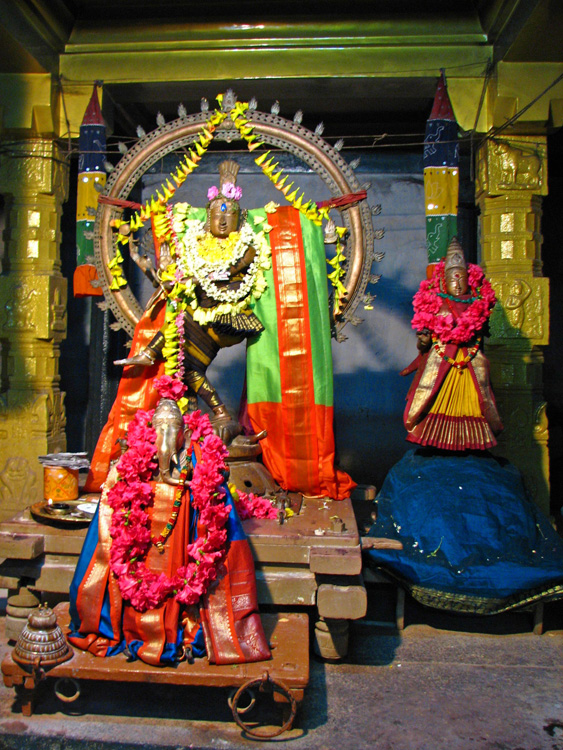 Three statues of Hindu gods garlanded with flowers are seen inside a temple setting.