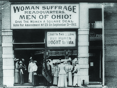A photograph shows women suffragists standing outside a building. The sign above them reads 